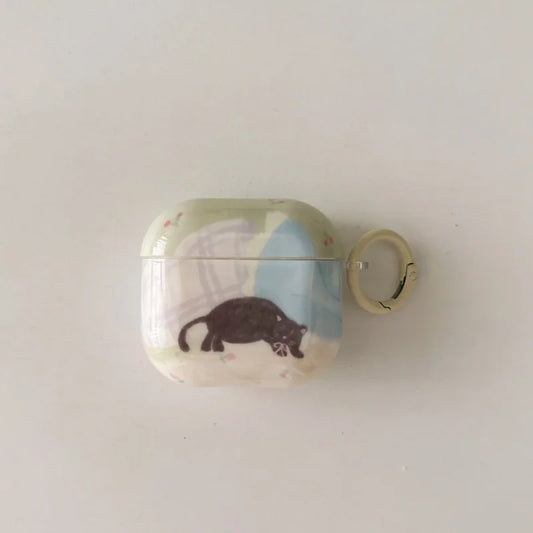 A wireless earbud charging case with a light green and blue abstract pattern, featuring a black cat illustration and a small beige key ring attached to the side, set against a plain off-white background.