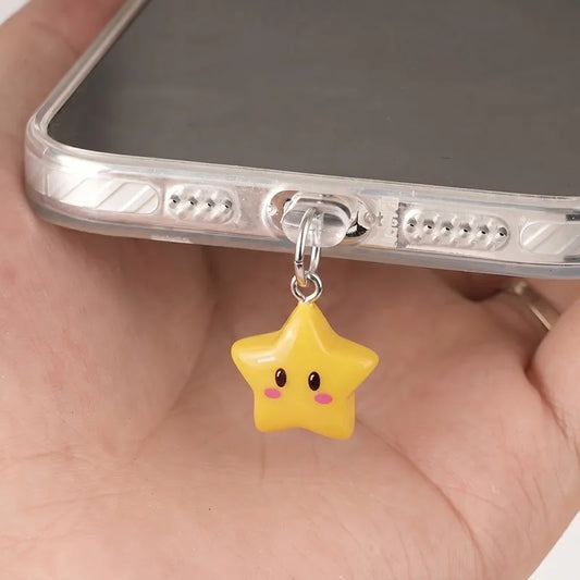 A yellow star-shaped phone charm with a cute face hangs from a silver metal ring on a clear phone case, held in a person's hand.
