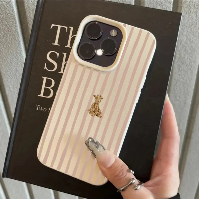 A hand holding a phone case with vertical pink and cream stripes, featuring a small gold giraffe illustration and a raised camera bumper, positioned over a black book.