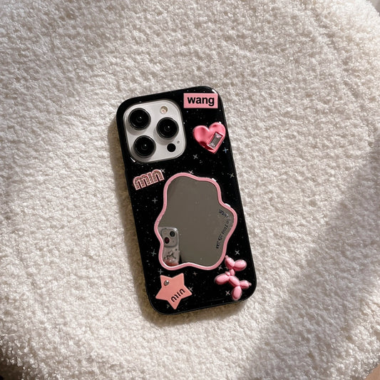 A black phone case with pink and silver custom charms, including a heart-shaped mirror, a dog-shaped charm, and the names 'wang' and 'min', shown on a white textured blanket.