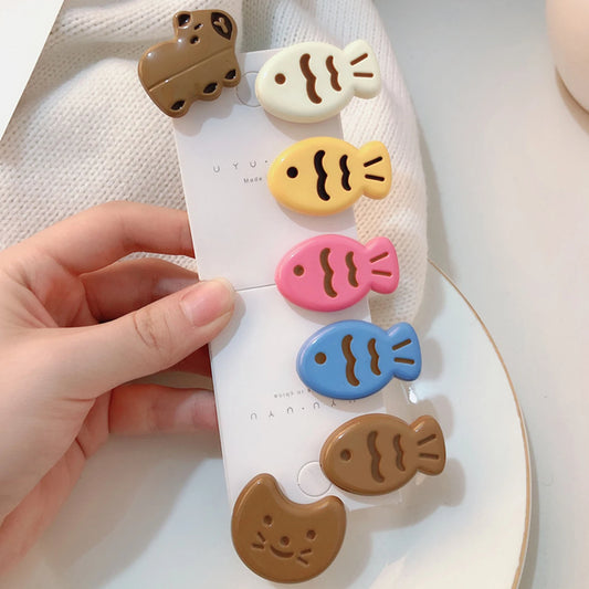 A hand holds a white card displaying five cute fish-shaped hair clips in pastel colors: brown, cream, yellow, pink, and blue. The clips have a glossy finish and are decorated with simple black line details.