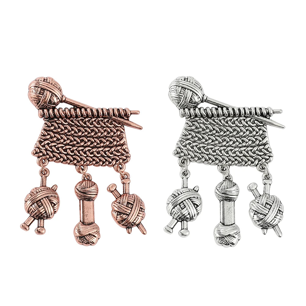 Two knitted scarf-shaped brooches with dangling yarn and knitting needle charms, shown in rose gold and silver metal finishes on a plain white background.