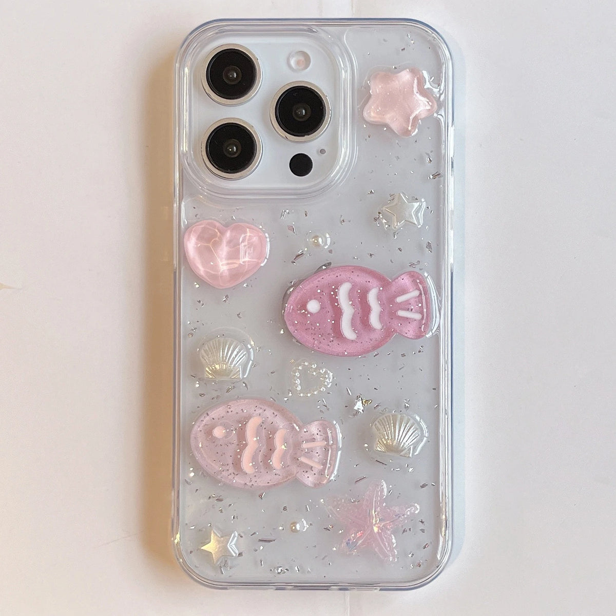 A clear phone case decorated with pink 3D fish, hearts, starfish, and shell designs, filled with silver glitter, shown on a white phone against a light background.
