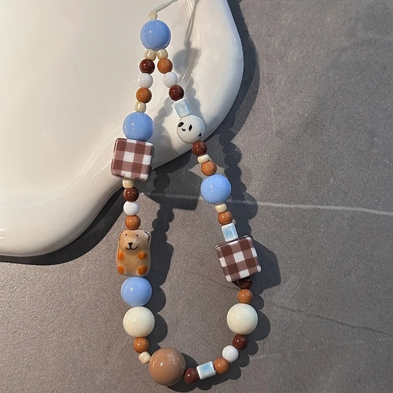 A beaded phone strap with light blue, brown, and cream beads, featuring a small bear charm and checkered square beads, displayed on a gray surface next to a white ceramic object.