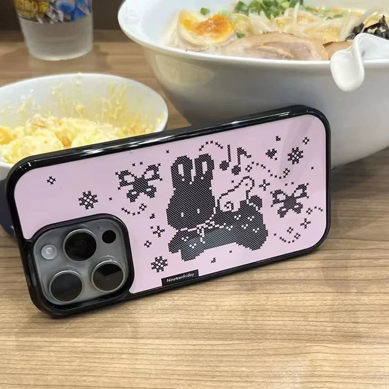 A pink phone case with a black pixelated bunny graphic and black bumper edges, shown resting on a wooden table with bowls of food in the background.