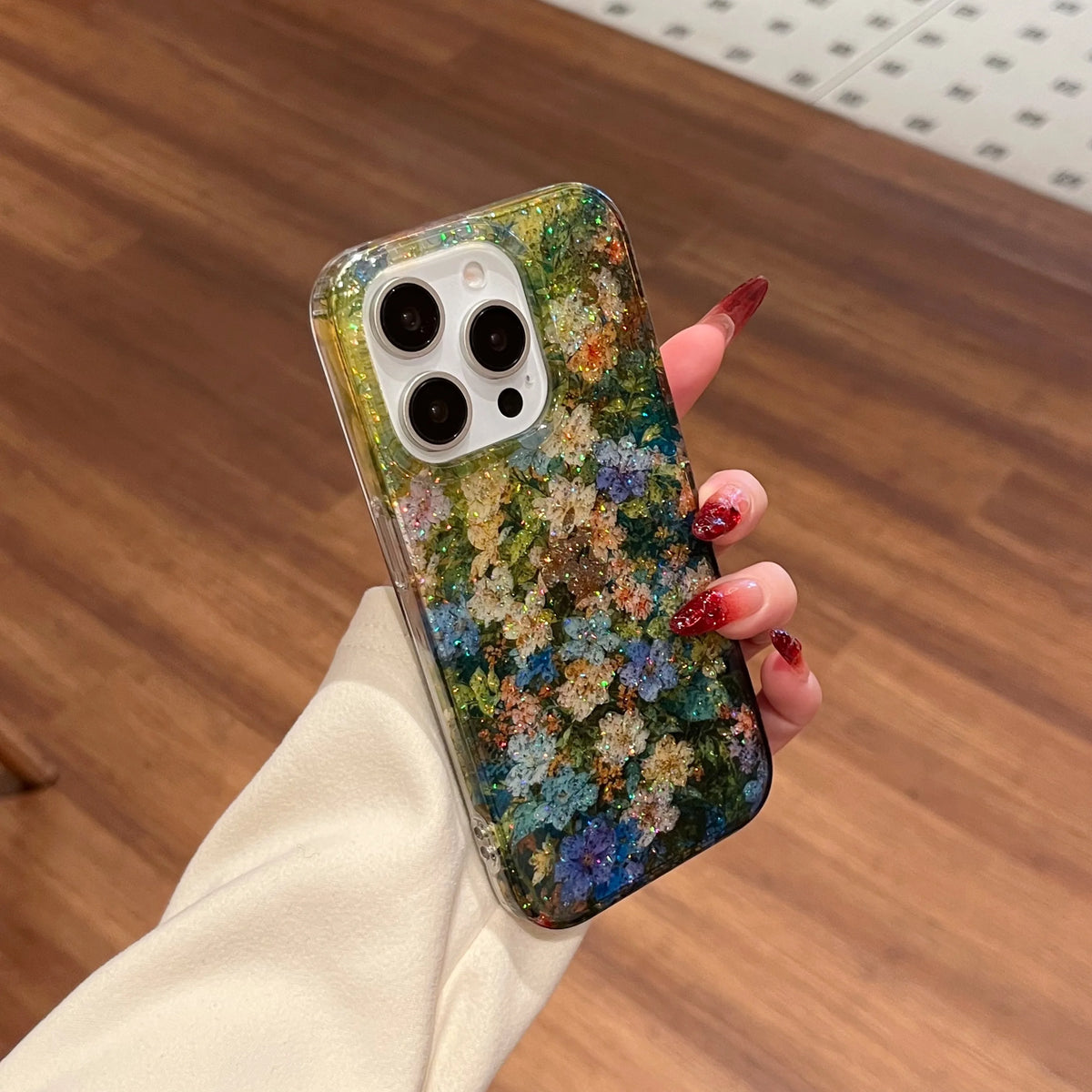 A hand with red glittery nails holds a clear iPhone case decorated with a dense, colorful pressed floral design in green, blue, yellow, and white. The background shows a warm wooden floor.