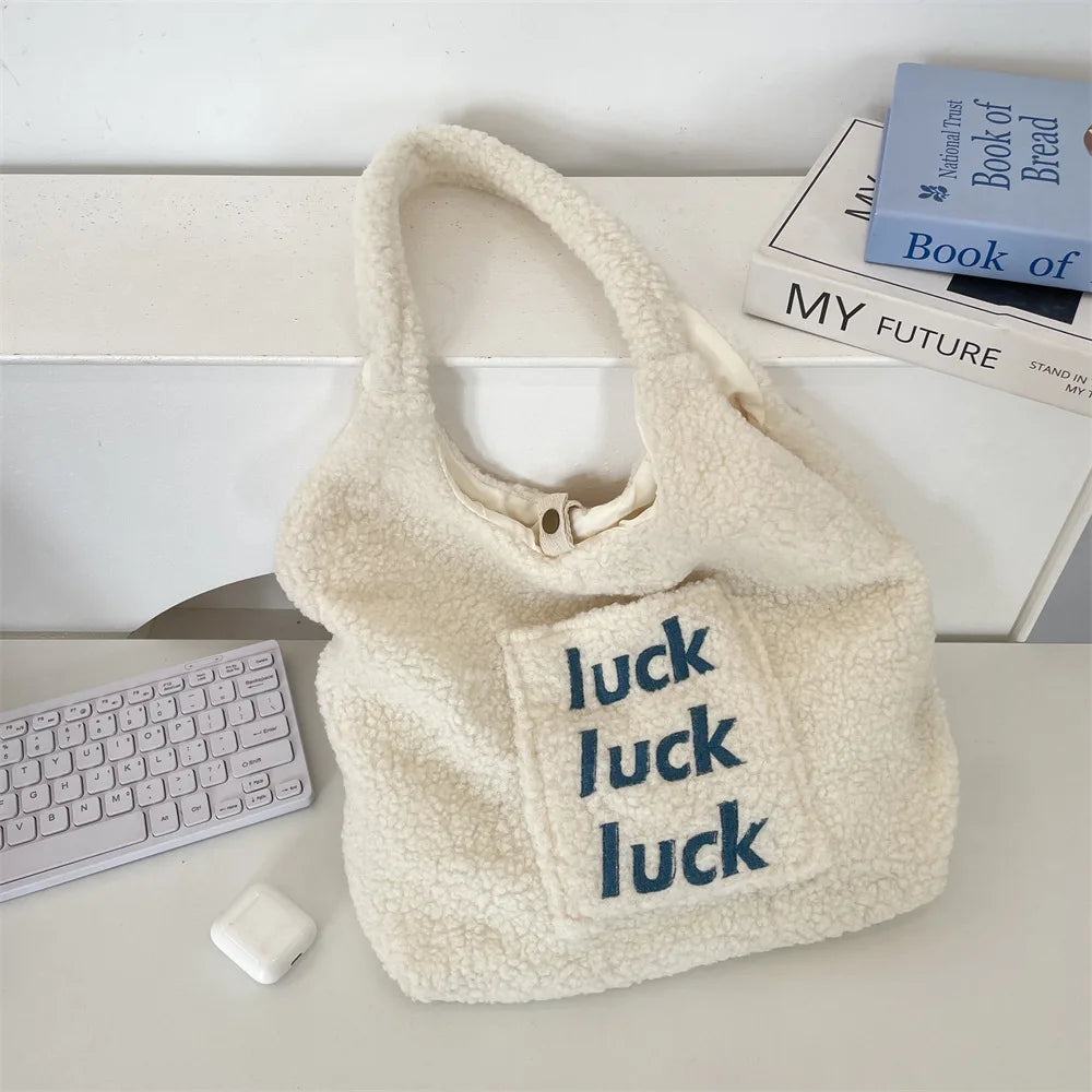 Cream-colored sherpa fleece shoulder bag with 'luck luck luck' embroidered in navy blue on the front pocket, styled on a white desk with a keyboard, earbuds case, and books in the background.