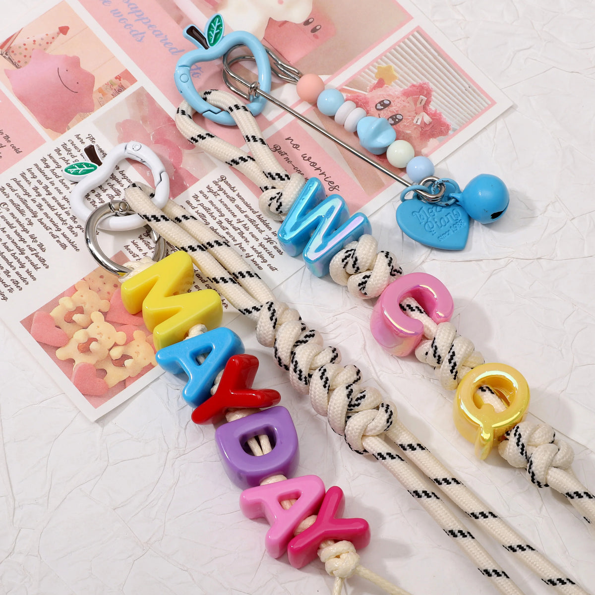 Three handmade rope letter keychains spelling 'WAKA' in blue, 'MAYA' in multicolor, and 'WAKO' in pink, decorated with colorful beads, charms, and metal keyrings, arranged on a textured white surface with printed cards in the background.