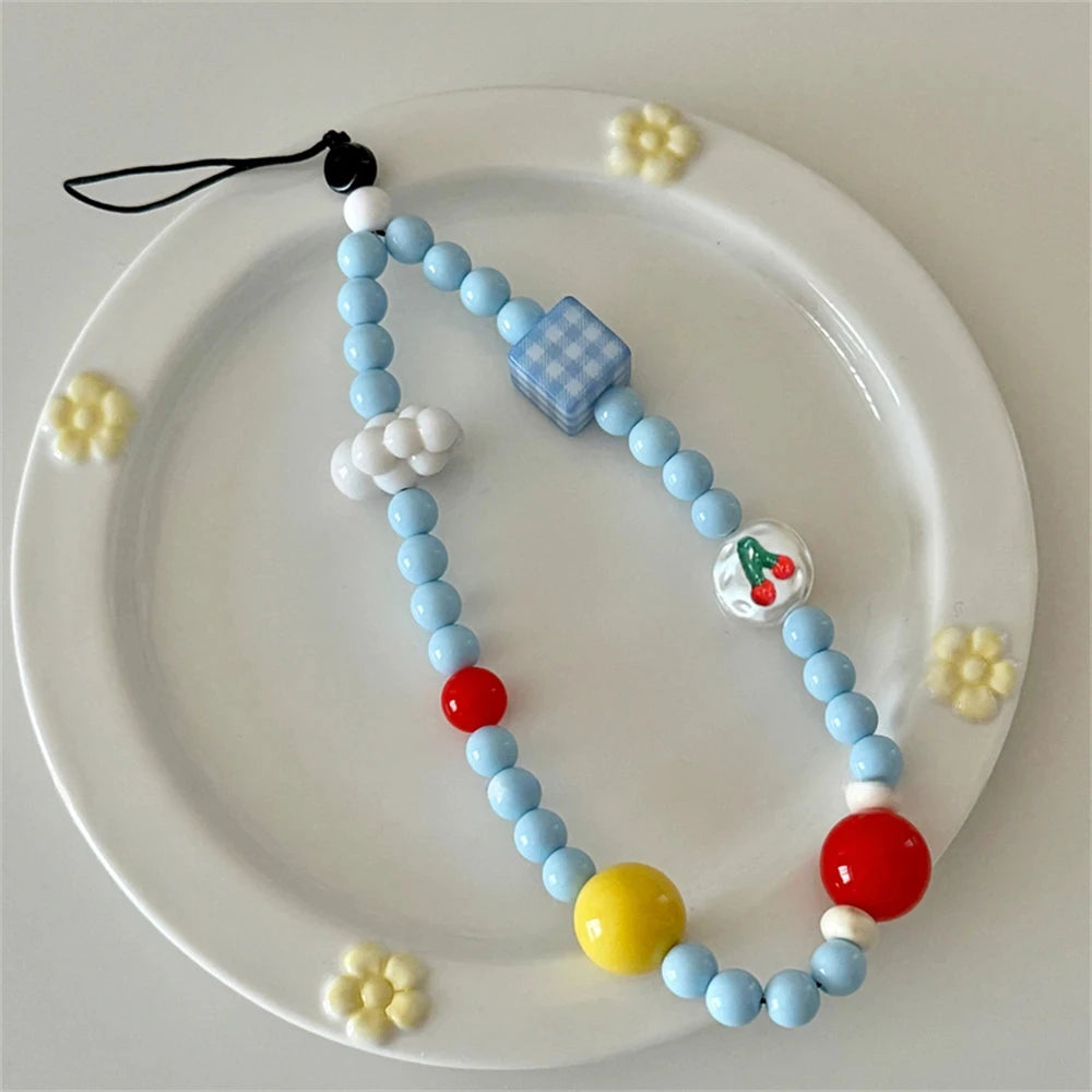 A light blue beaded phone strap with decorative charms, including a blue checkered cube, white clouds, a cherry bead, and red and yellow accent beads, displayed on a white ceramic plate with small yellow flower decorations.
