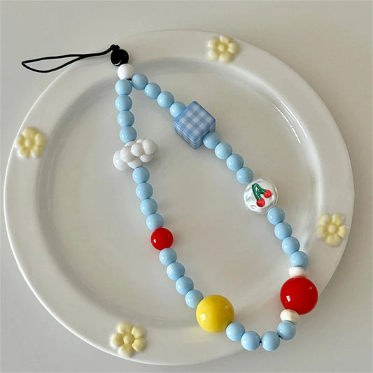 A light blue beaded phone strap with decorative charms, including a blue checkered cube, white clouds, a cherry bead, and red and yellow accent beads, displayed on a white ceramic plate with small yellow flower decorations.