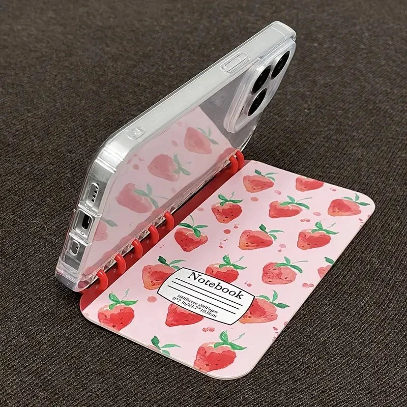 A clear smartphone case with a pink strawberry-patterned inner notebook cover, shown open on a dark textured fabric background.