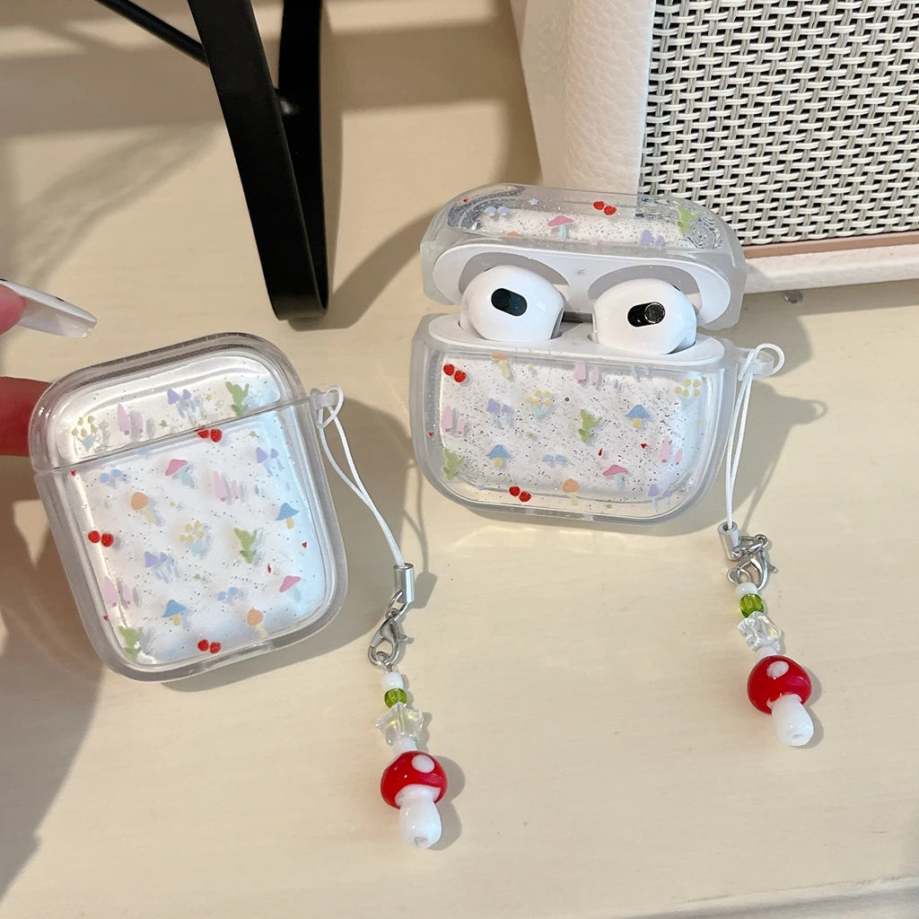 Clear AirPods case with a delicate multicolor floral print and glitter, shown with matching silicone earbud covers and mushroom-shaped charm straps on a light beige tabletop.