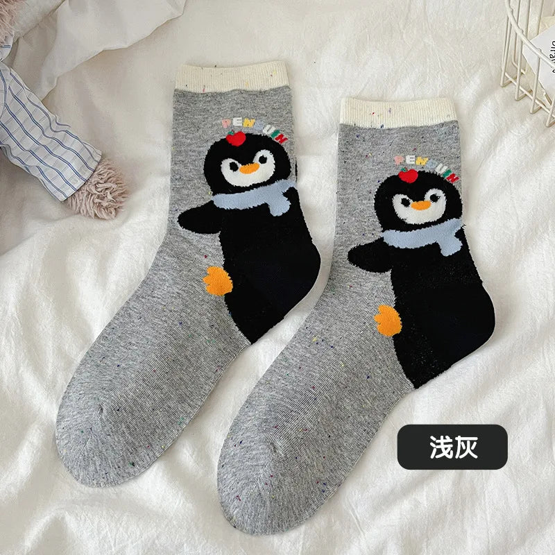 A pair of light gray heathered ankle socks with a cartoon penguin design on the side. The penguin is black with a light blue scarf, orange beak and feet, and a small multicolor hat.