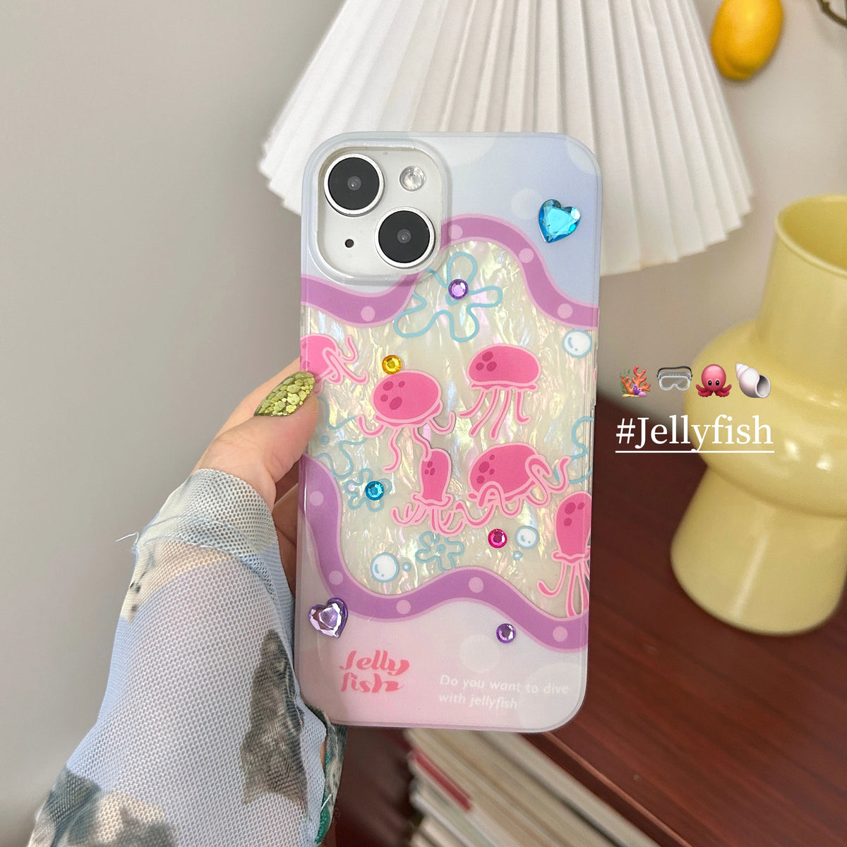 A hand holds a phone case decorated with pink jellyfish, blue heart charms, and iridescent glitter. The case features a pastel gradient background transitioning from light blue to pink, with a wavy purple border.
