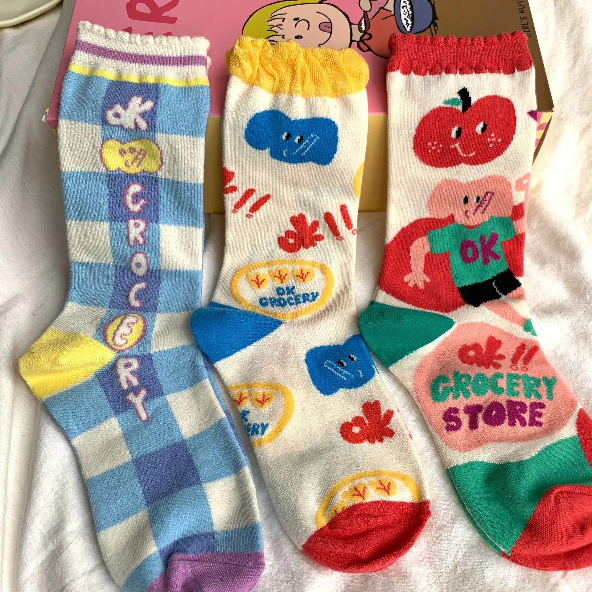 Three colorful novelty socks laid flat on a white fabric surface, featuring cartoon grocery-themed prints including a striped blue-and-white pattern, blue cloud motifs, a red apple character, and a pink grocery store sign.