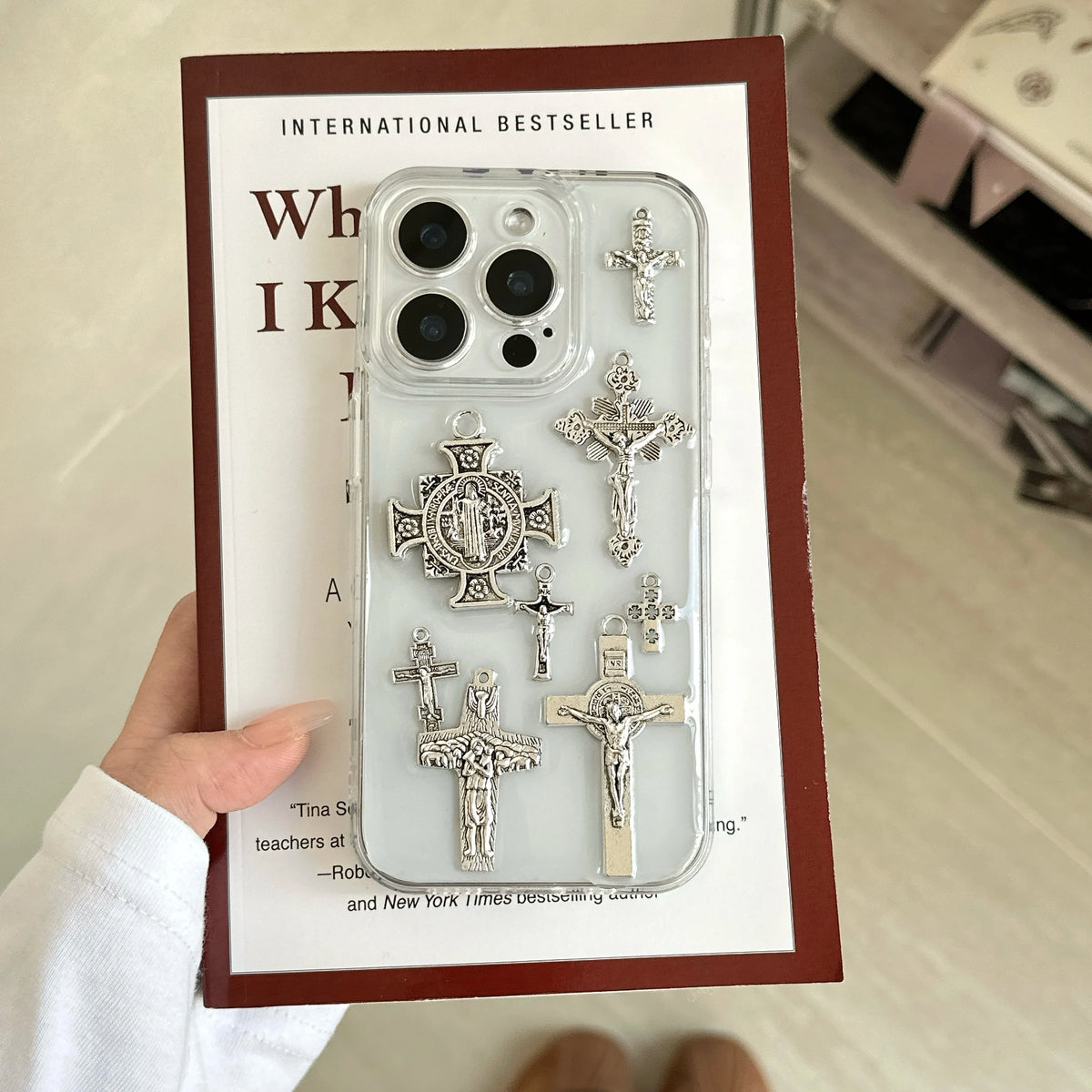 A hand holds a clear phone case decorated with multiple silver-tone religious cross and crucifix charms, presented on a white backing card with a dark red border.