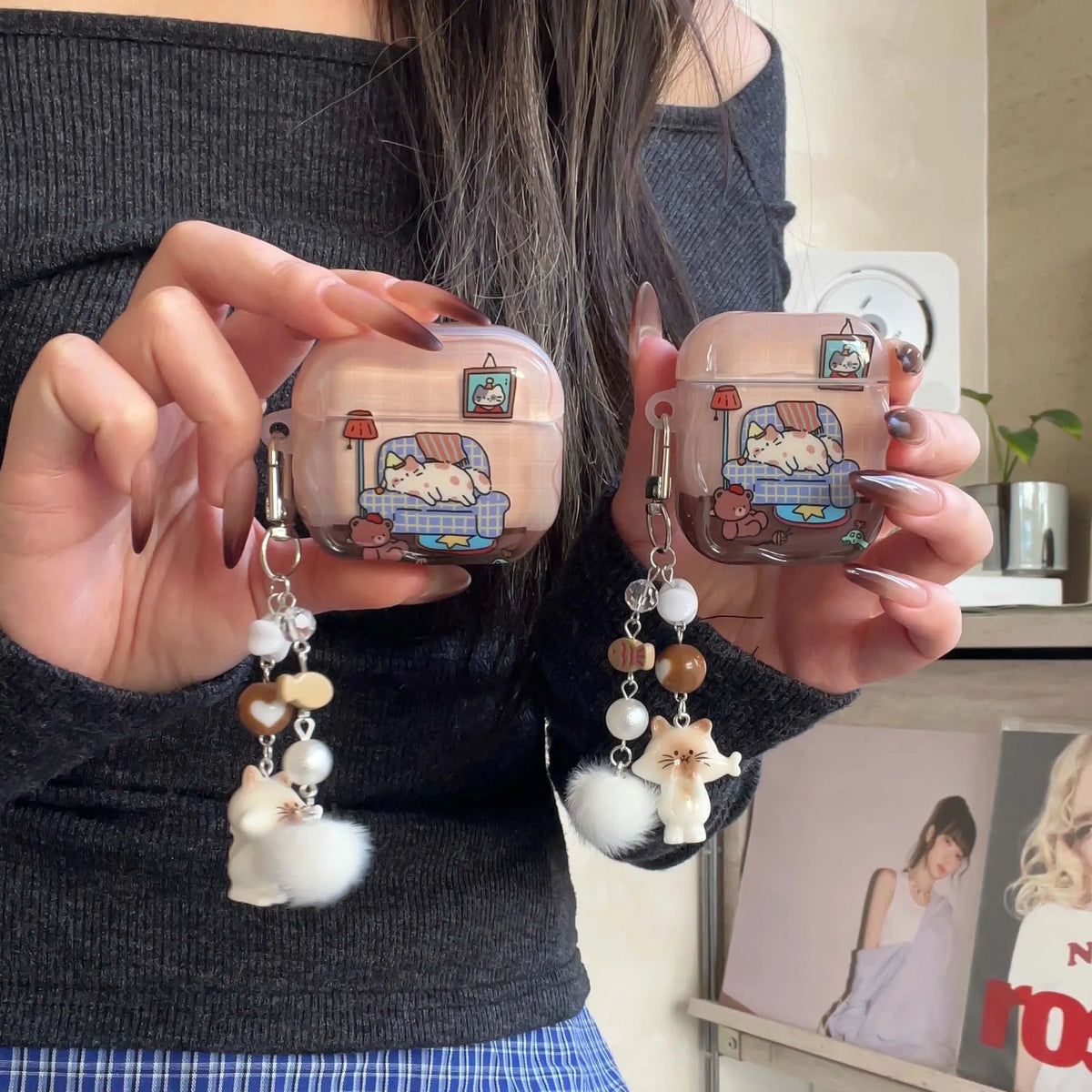 A person holds two transparent pink AirPods cases decorated with a cute cartoon cat sitting on a blue sofa charm, each with dangling beaded chains and white cat poms, against an indoor background.
