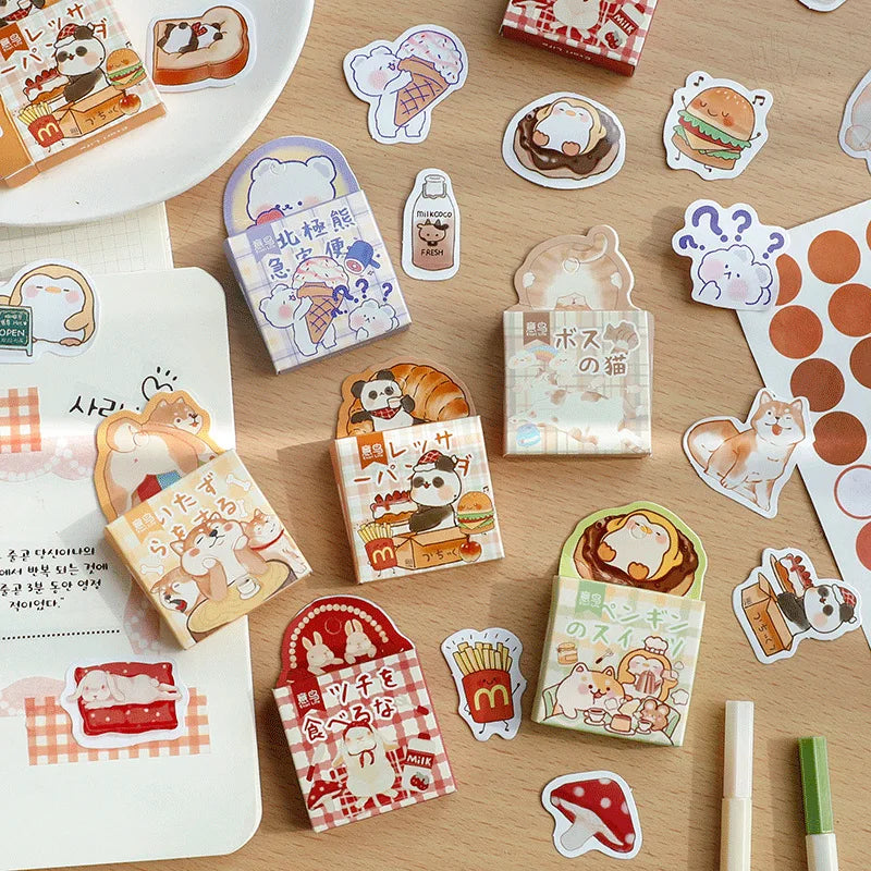 A styled flat-lay of cute cartoon sticker sheets and individual sticker cutouts on a light wood desk. The stickers feature various food characters, including a panda with a coffee cup, a croissant, a hamburger, french fries, and a cat, alongside Japanese text and checkered patterns.