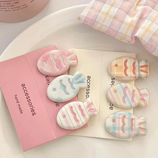 Four pastel fish-shaped hair clips arranged on a white plate with a pink card and a folded plaid cloth nearby. The clips are translucent and decorated with soft pink, blue, yellow, and white stripes and plaid patterns.