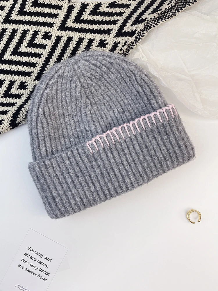 A gray ribbed knit beanie with a folded cuff and a row of light pink embroidered details, styled on a white surface with a black-and-white patterned textile, a white plastic bag, and a small gold ring.