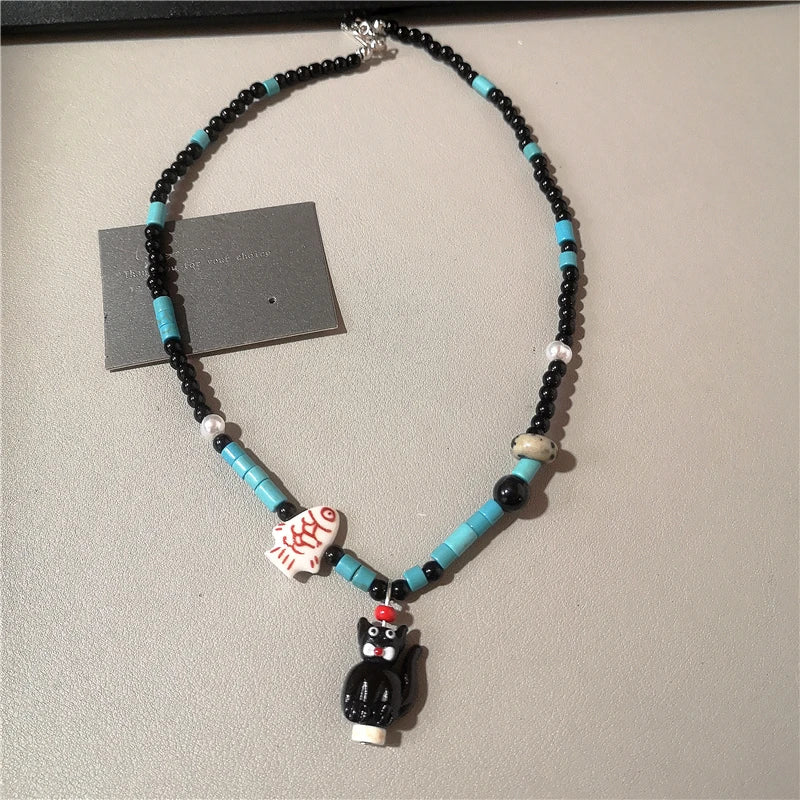 A handmade beaded necklace featuring turquoise and black beads, a white fish-shaped charm with red text, and a black cat pendant, displayed on a neutral beige surface.