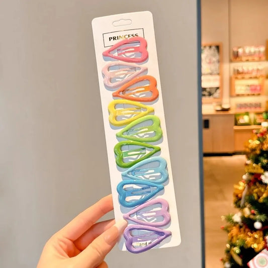 A hand holds a white card displaying eight pastel-colored heart-shaped hair clips in pink, blue, orange, yellow, green, and purple. The card is labeled 'PRINCESS' at the top, and the background shows a blurred indoor setting with a Christmas tree.