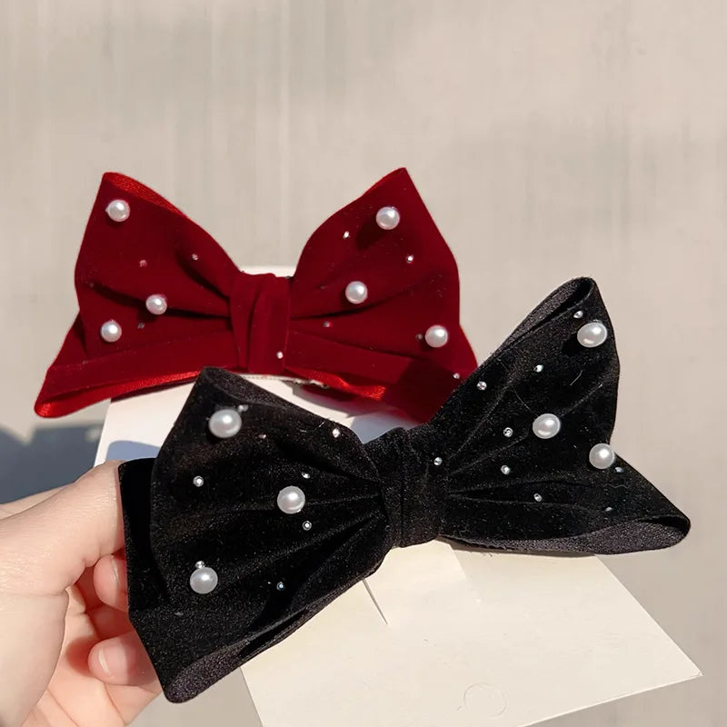Two velvet bow hair clips, one red and one black, both decorated with small white pearls and rhinestones, shown with a hand holding the black one against a light neutral background.