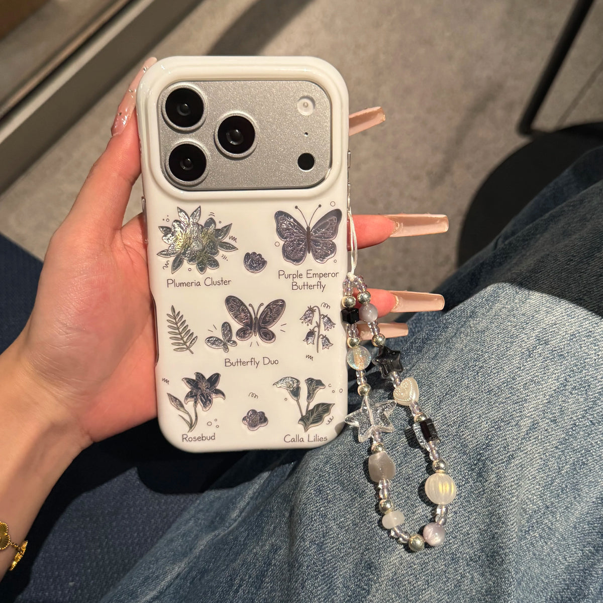 A hand holds a white phone case decorated with vintage-style botanical and butterfly illustrations, including labels like 'Plumeria Cluster' and 'Purple Emperor Butterfly.' The case has a matching beaded wrist strap.