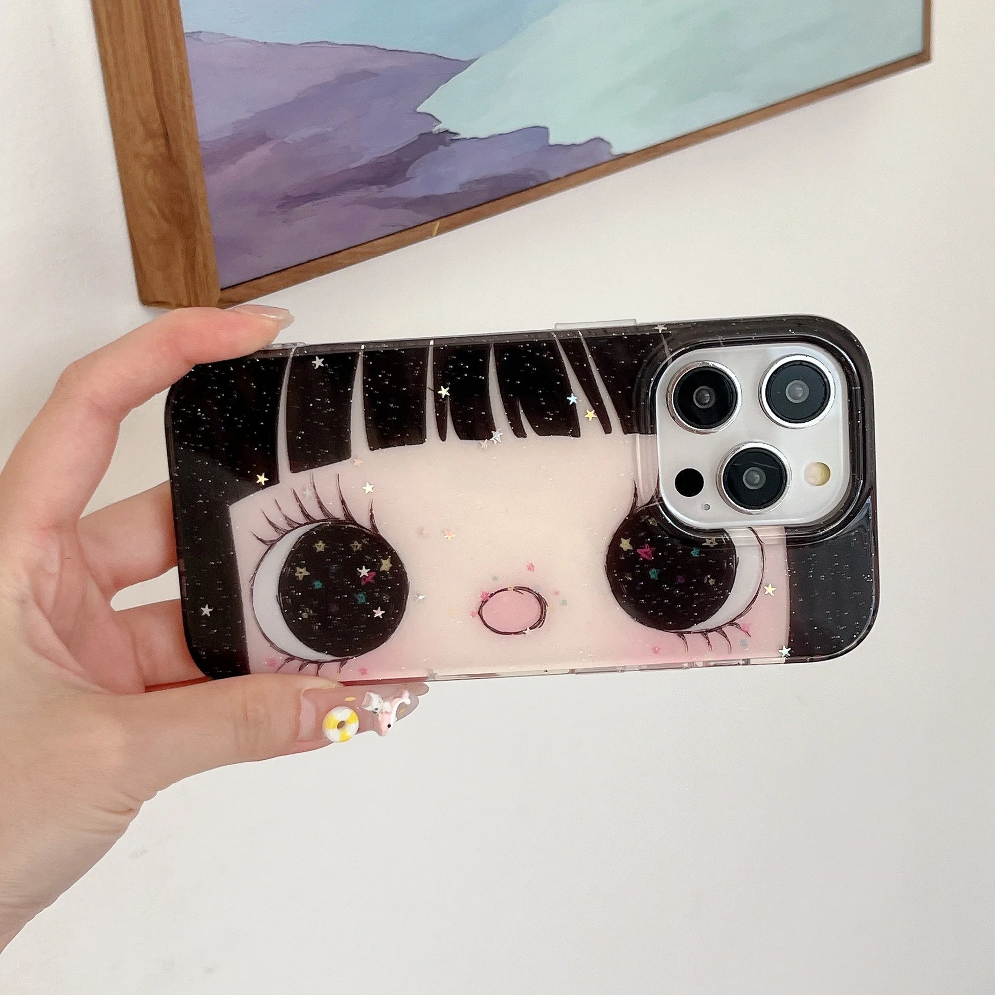 Phone case with cartoon face design held by a hand, with a painting in the background.