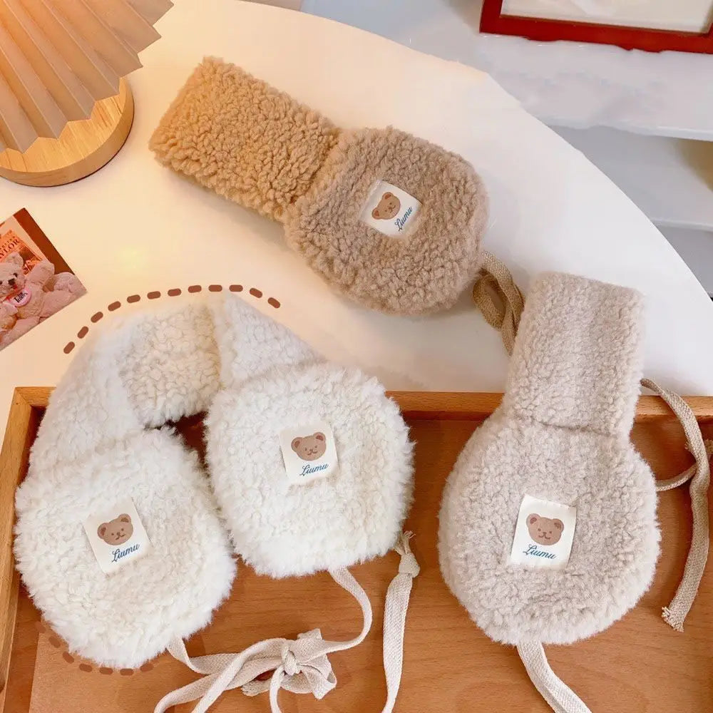 Three pairs of fuzzy sheepskin earmuffs in cream, beige, and light brown, arranged on a wooden tray and a white table. Each pair has a small white patch with a teddy bear face and the brand name 'Bourou'.