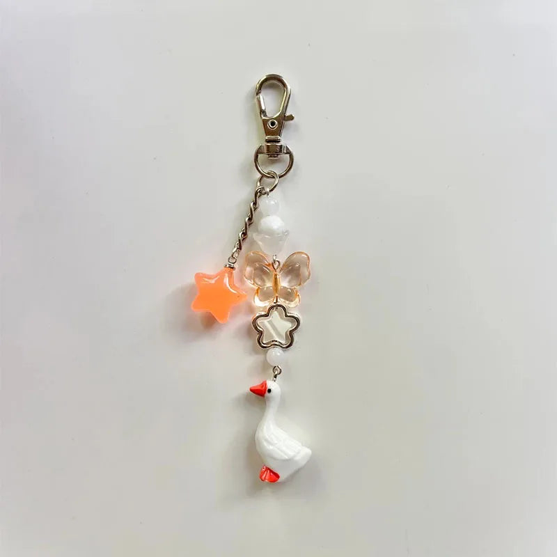 A charm keychain with a gold-tone lobster clasp, featuring a white goose charm with orange beak and feet, a white bead, a translucent butterfly charm, a white flower charm, and an orange star charm, all on a plain light background.