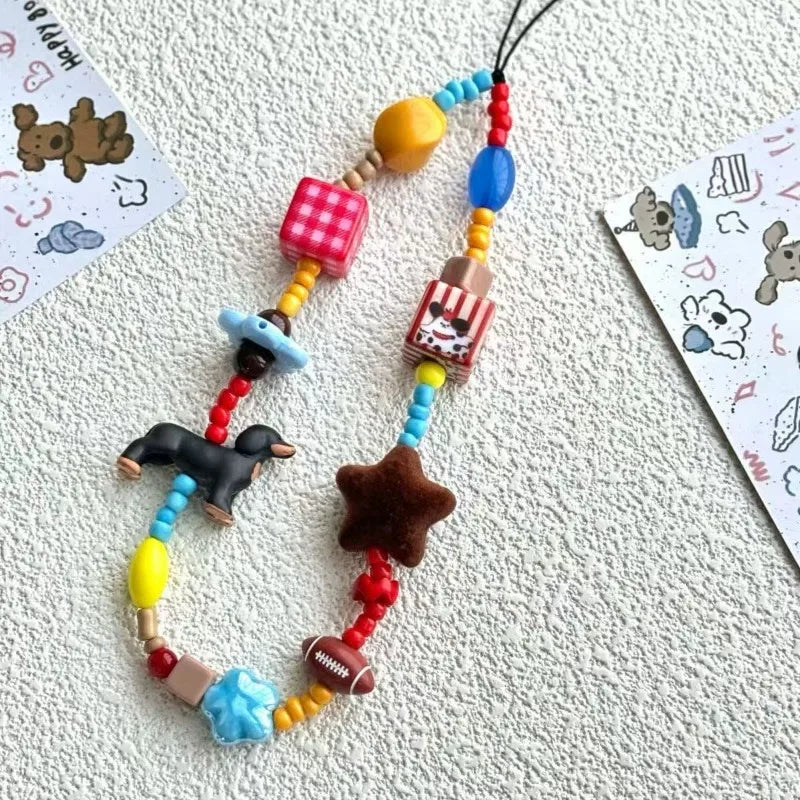 A colorful beaded phone strap or charm laid on a textured white surface, featuring small toy-like beads and charms including a dachshund figure, a star, a red-and-white striped popcorn bucket, a pink gingham cube, and blue and yellow beads.