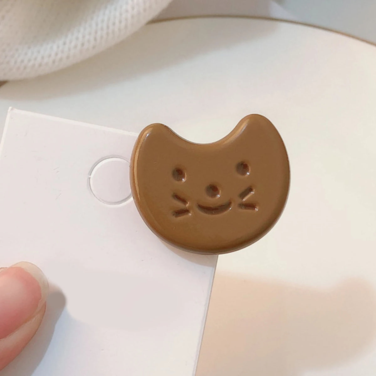 A small brown crescent-shaped accessory with a simple cat-face design, shown resting on a white card with a finger visible at the lower left.