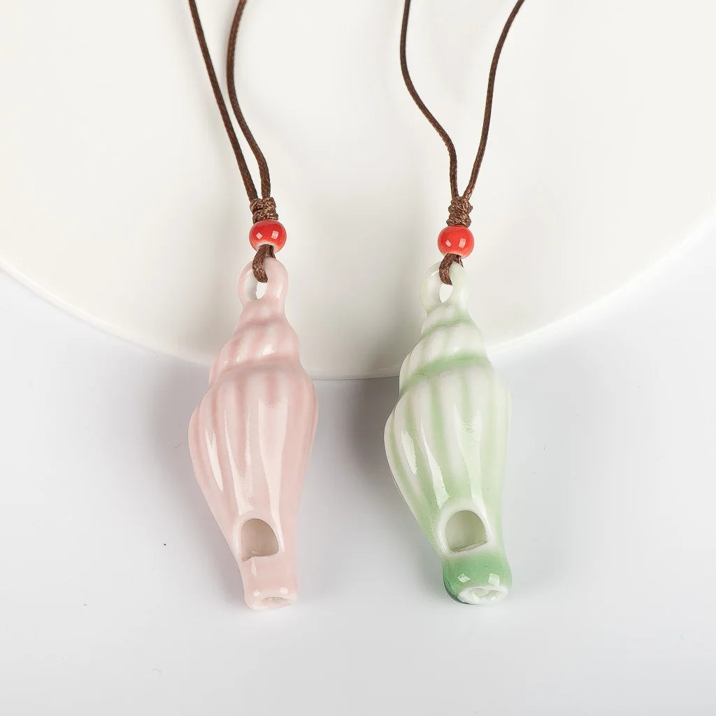 Two seashell-shaped pendant necklaces on brown cords, one in light pink and one in light green, each accented with a small red bead, displayed against a white background.