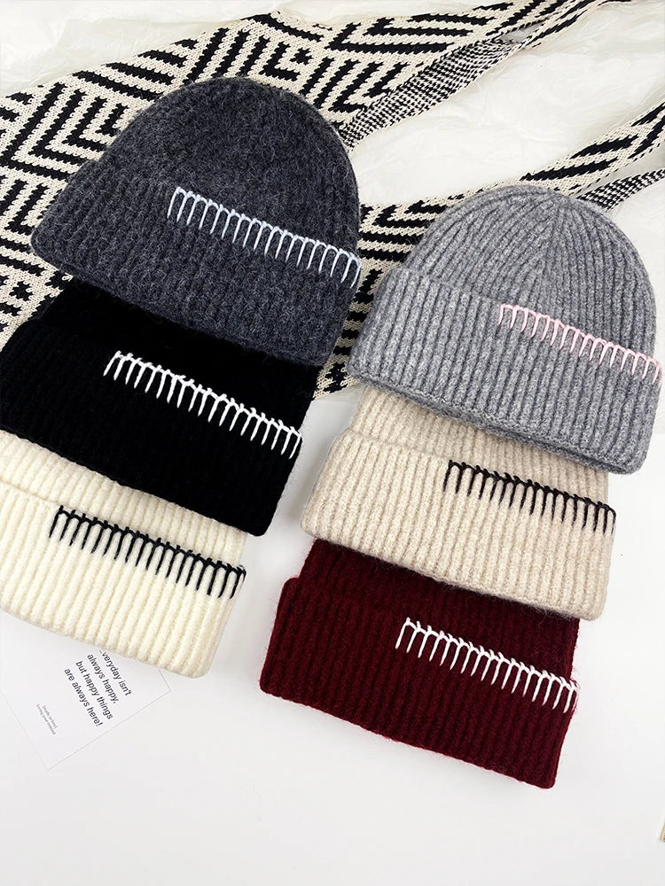 Four ribbed knit beanies arranged in two columns on a white surface with a black-and-white patterned textile behind them. The beanies are stacked by color: dark gray on top, then black, then cream; next to them are light gray, cream, and burgundy, each with contrasting embroidered stripe details.