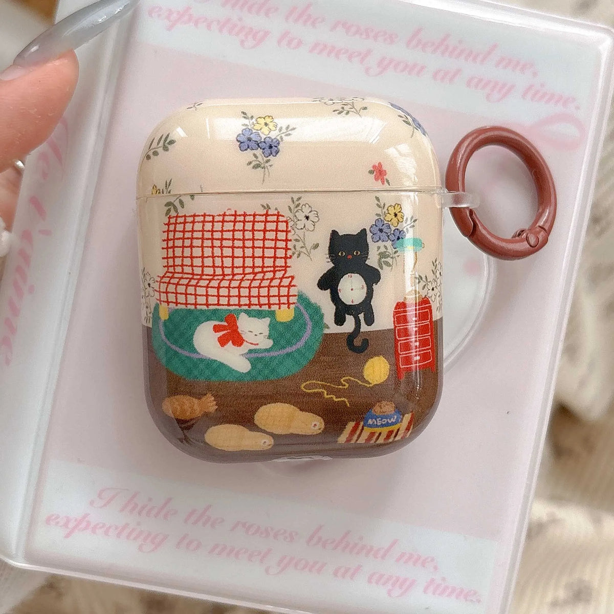 A clear protective case for an AirPods charging case, featuring a cute Japanese-style illustration of a black cat and a white cat with floral patterns, resting on a printed card with a clear plastic ring.