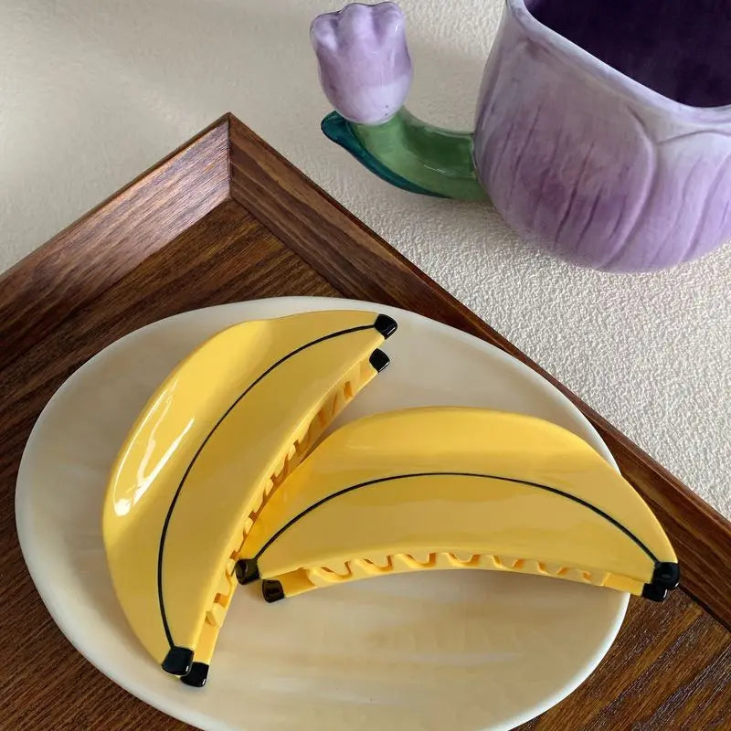 Two yellow hair claw clips shaped like bananas, displayed on a white plate on a wooden tray.