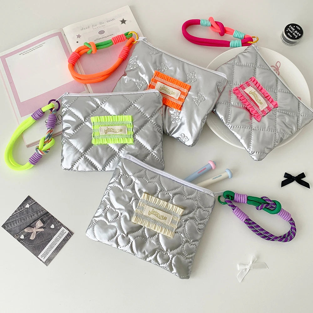 Four quilted silver coin pouches with heart patterns, each featuring a different colored braided wrist strap (green, orange, pink, purple) and a decorative fabric label. The pouches are arranged on a white surface with various stationery and small accessories.