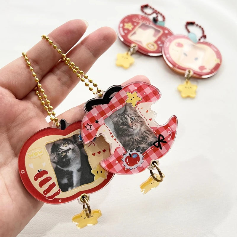 A hand holds a set of acrylic keychains shaped like a red-and-white checkered apple and apple core, both featuring a photo of a gray cat in the center. The keychains are decorated with small stars, hearts, and a red bow, and hang from gold-tone chains with small yellow charms.