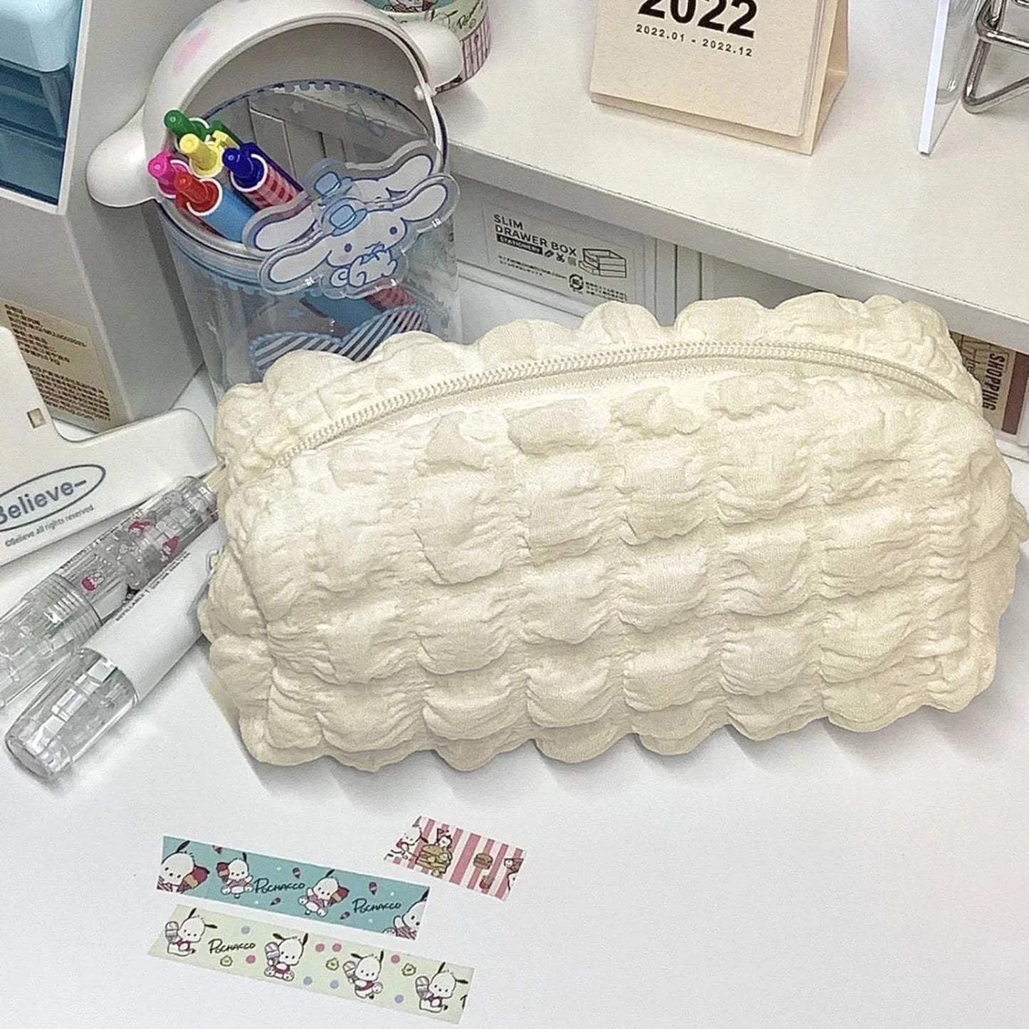 A cream-colored, textured bubble-patterned zippered pencil case or cosmetic pouch, displayed on a white desk alongside stationery items including washi tape, pens, and a Cinnamoroll-themed cup.