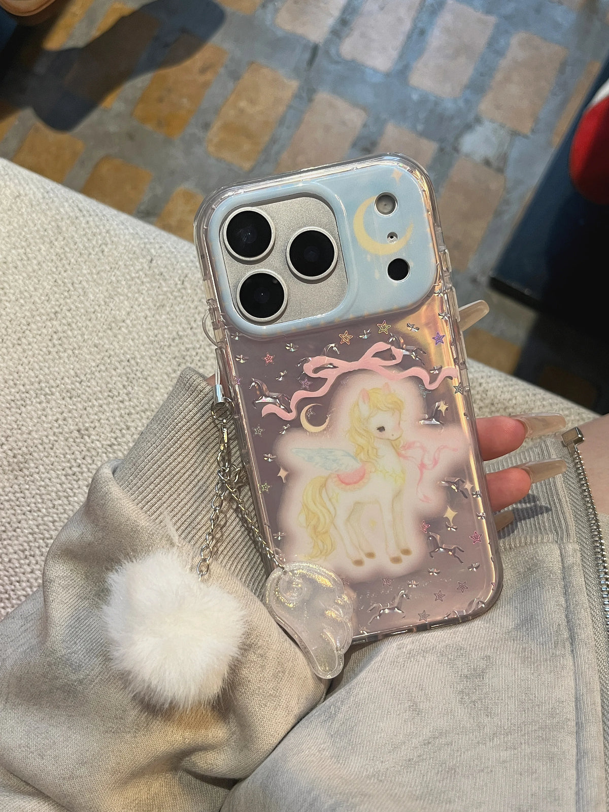 A clear phone case with a pastel unicorn illustration, pink stars, and a small moon detail, shown on a phone held in a hand. The case is resting on a light gray fabric surface with a patterned floor in the background.