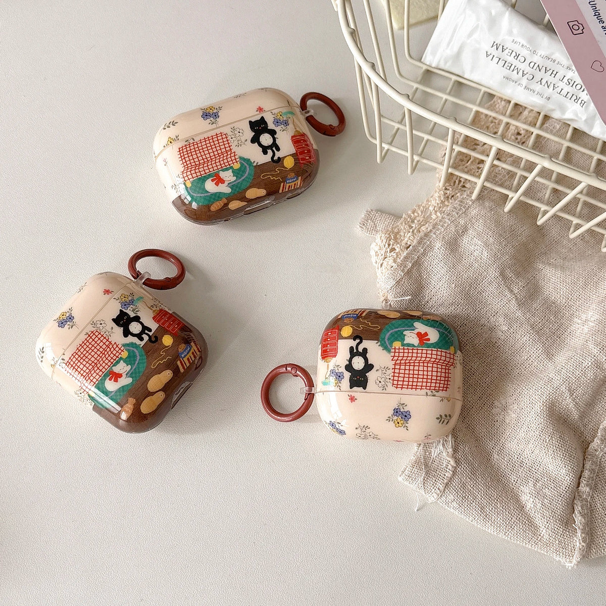 Three decorative AirPods cases with red carrying loops are arranged on a light surface beside a linen cloth and a wire basket containing skincare items. The cases have a cream background with a whimsical multicolor illustration featuring black cats, red-and-white checkered patterns, and small floral motifs.