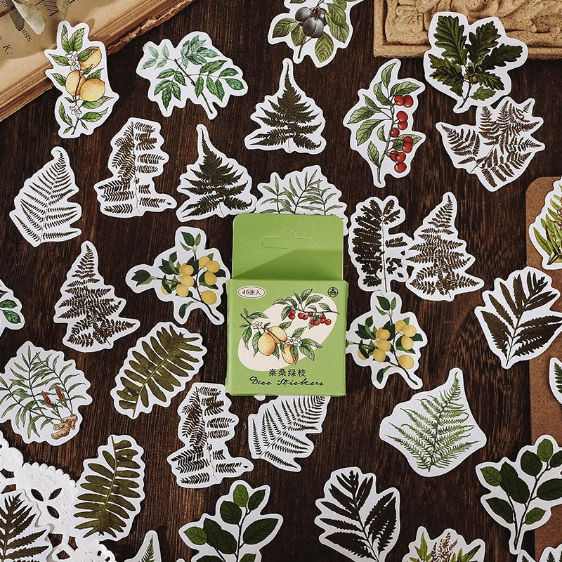 A set of botanical and fruit-themed die-cut stickers arranged on a dark wooden surface, with a small green sticker box in the center. The stickers feature illustrated ferns, leaves, berries, and fruit motifs.
