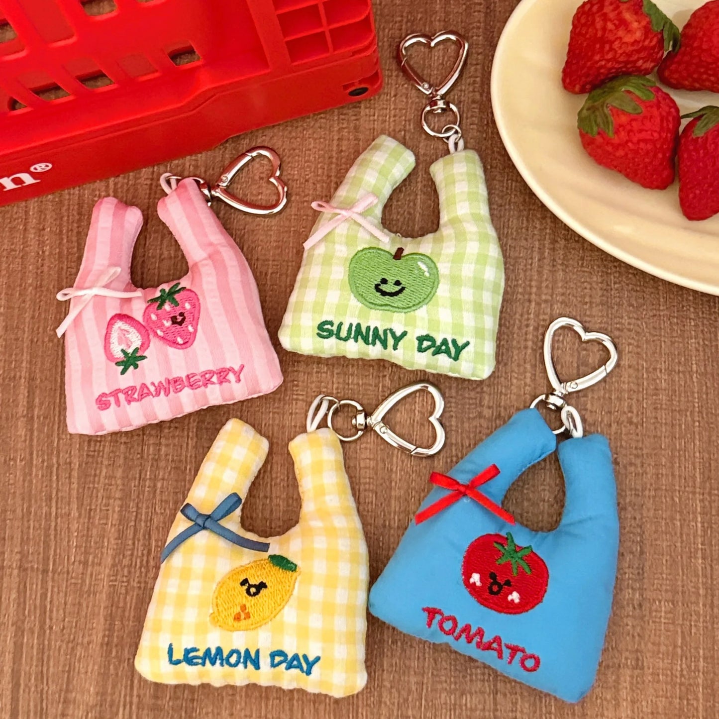 Four small fabric keychain bags shaped like shopping bags, each with a different fruit design and text: Strawberry, Sunny Day, Lemon Day, and Tomato. They are arranged on a brown textured surface with a red basket and strawberries in the background.