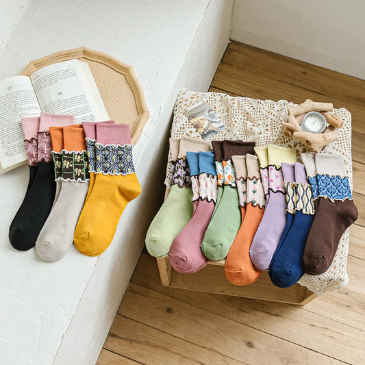 A styled flat-lay of multiple pairs of decorative ankle socks arranged on a lace-covered wooden box and leaning against a white wall beside an open book. The socks come in many colors and patterns, including black, cream, mustard yellow, pink, blue, green, orange, purple, and brown, with lace and floral cuffs.