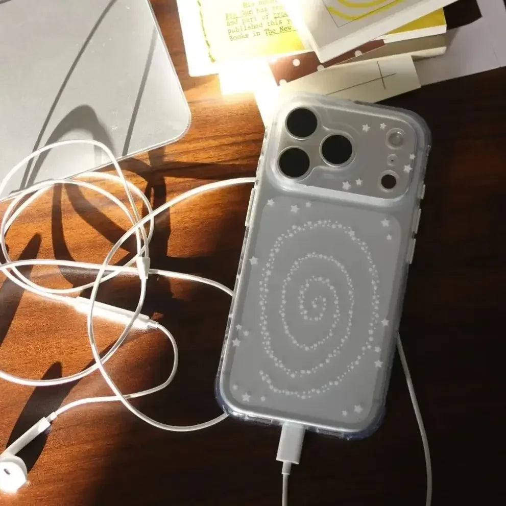 A clear phone case with a white spiral galaxy pattern and small star details, shown on a smartphone resting on a wooden desk with white earphones and cables nearby.
