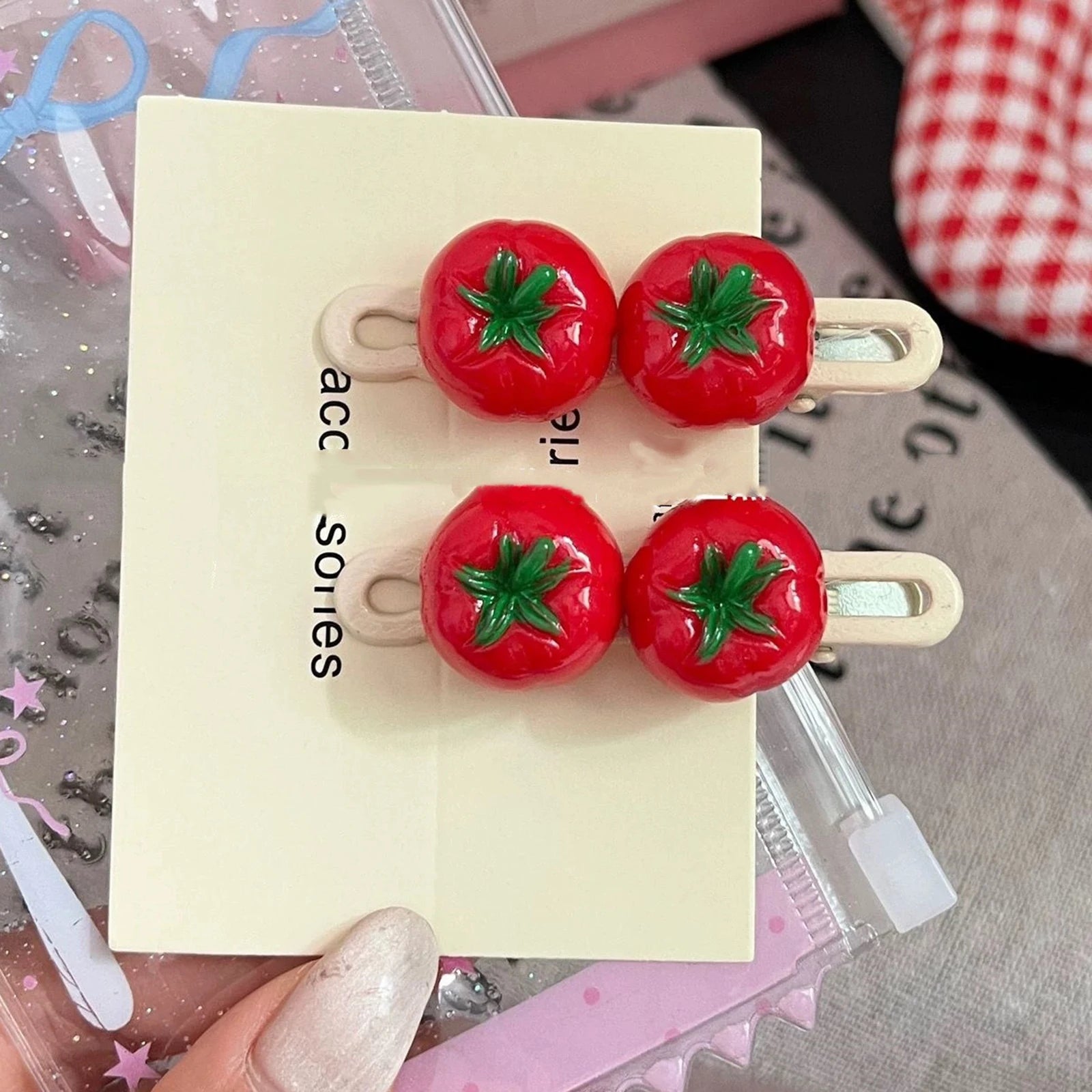 Four red tomato-shaped hair clips with green leafy tops, displayed on a cream-colored card held by a hand with glittery nail polish.