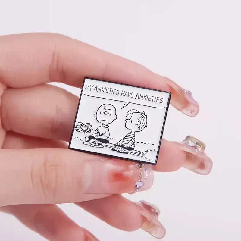 A hand with clear rhinestone nails holds a small rectangular enamel pin featuring a black and white cartoon of two people with the text 'MY ANXIETIES HAVE ANXIETIES'.