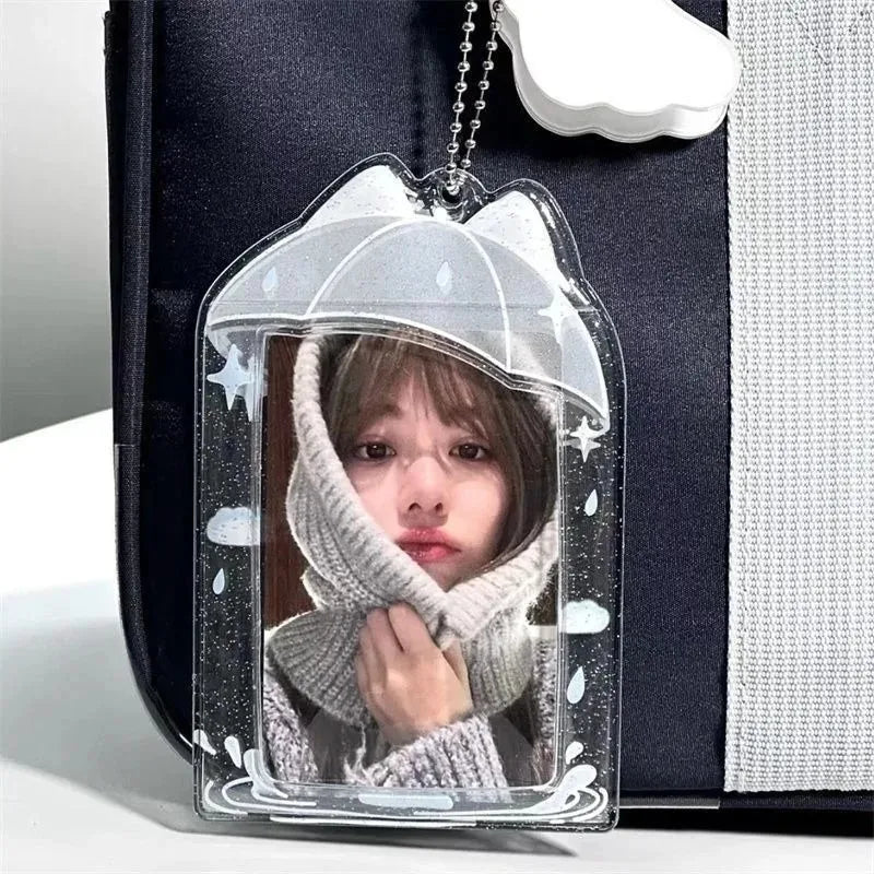 A clear acrylic keychain or bag charm featuring a printed photo of a girl in a white knit hood, framed by a clear umbrella design with white clouds and stars, shown hanging against a black and white bag.