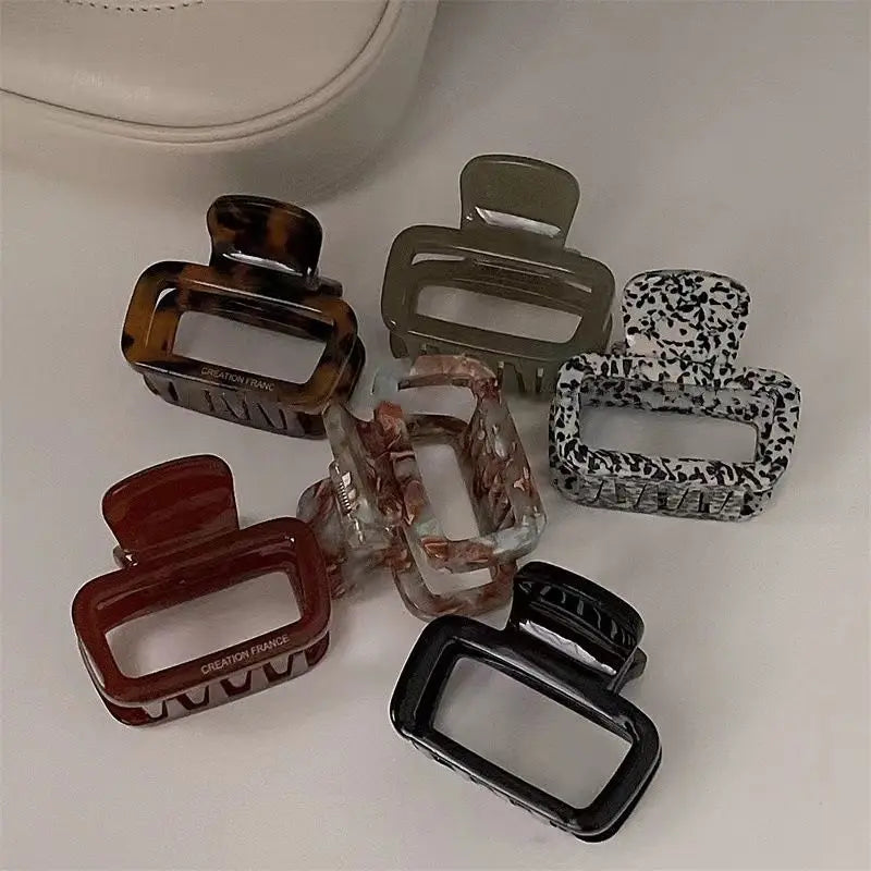 Six rectangular hair claw clips in assorted colors and patterns, including tortoiseshell, olive green, black-and-white speckled, burgundy, marbled pink, and solid black, arranged on a light beige surface.
